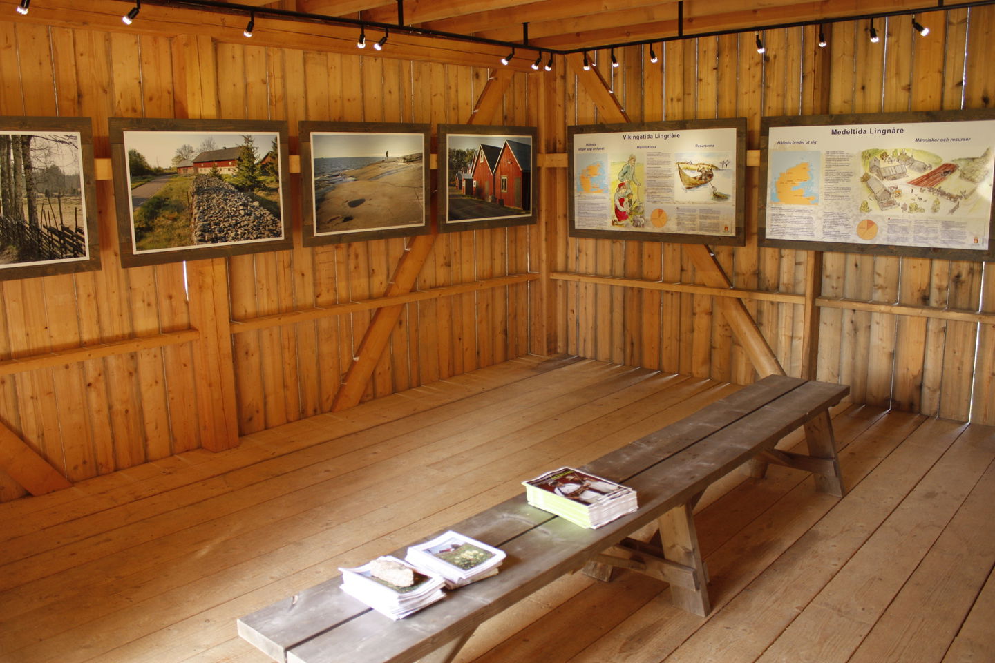 Mitt i ett rum står en sittbänk med tre högar av broschyrer på. På väggarna runtom hänger det tavlor.