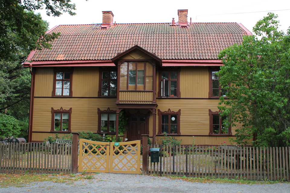 Finnstugan är ett tvåvåningshus med mörkgul träpanel, röda knutar och rött tegeltak.