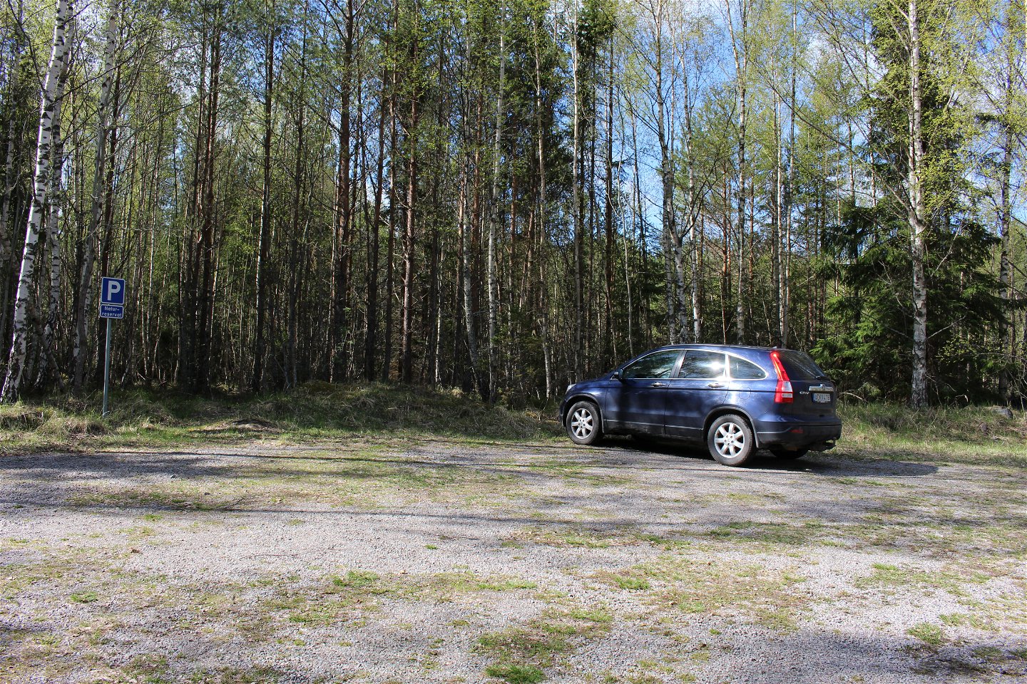 En bil står på en grusad parkeringsplats med skog runtom.