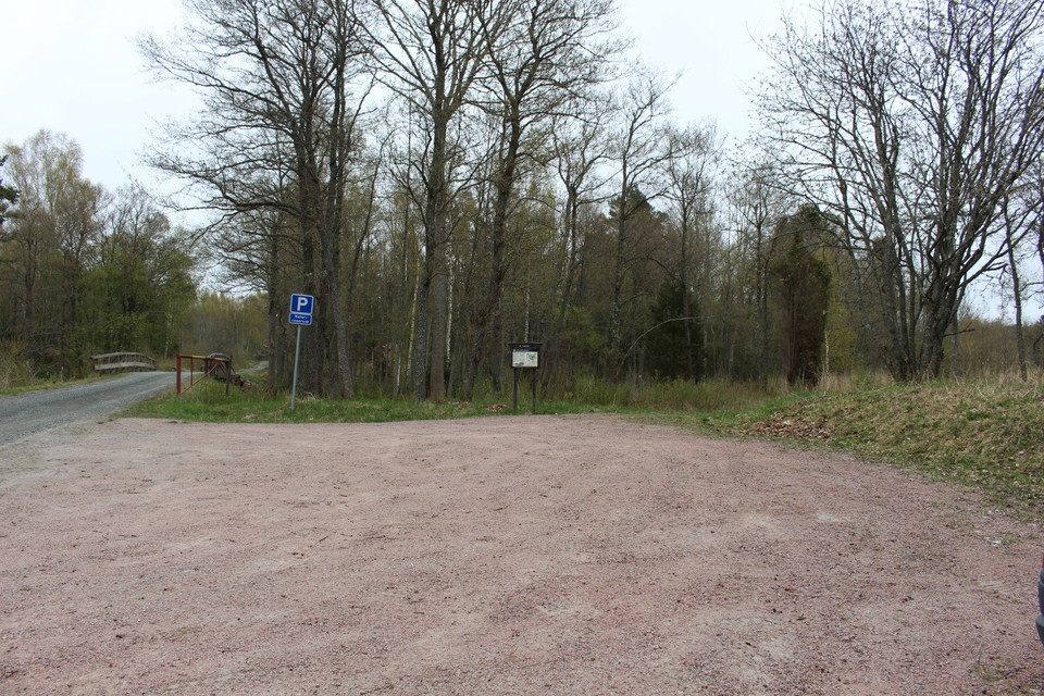En grusad parkeringsplats intill en mindre bilväg. Vid ena sidan finns parkeringsskylt och informationstavla. Parkeringen angränsar till en skogsdunge.