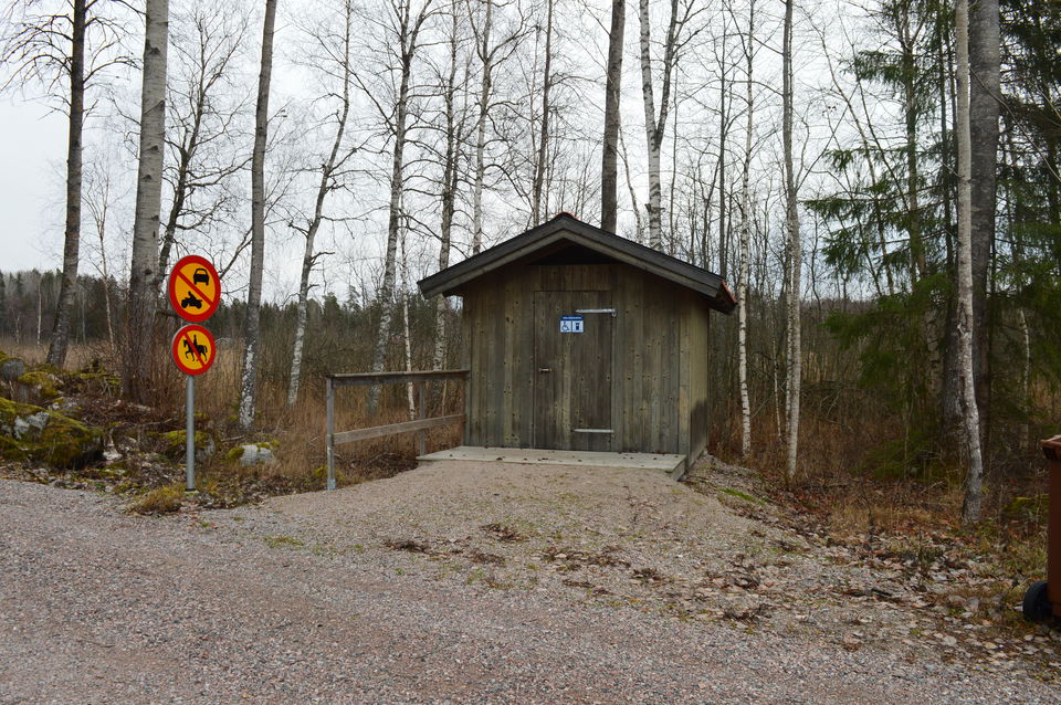 En torrtoalett med ett handräcke framför står vid en jämn grusad yta. Nära handräcket finns en vägskylt och bakom torrtoaletten växer träd.