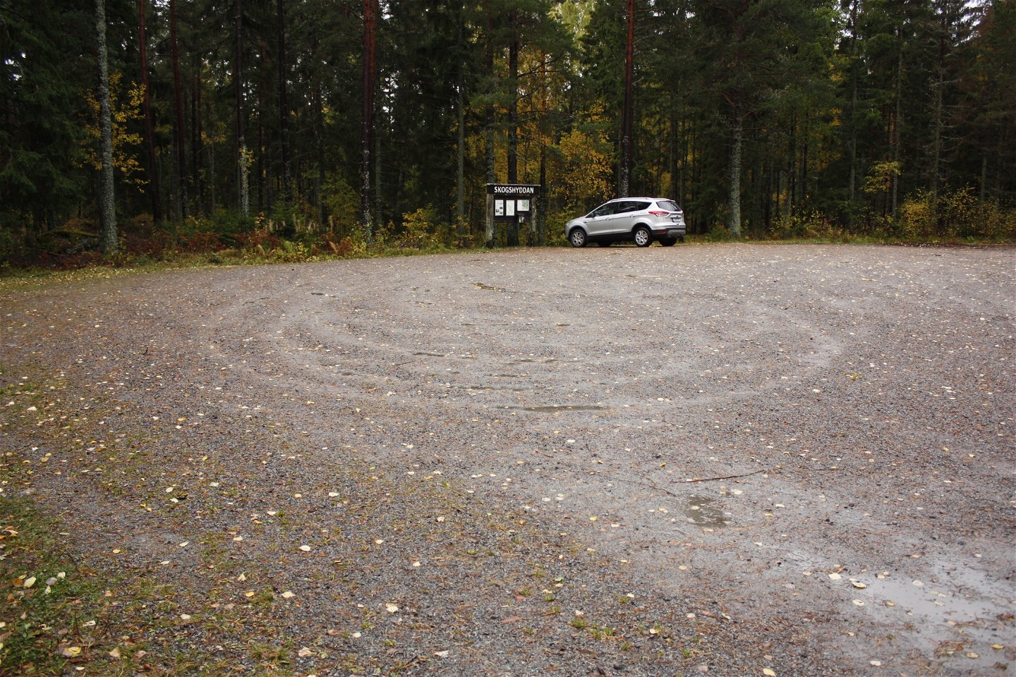 En bil står parkerad på en grusad parkeringsplats vid en skog. Framför bilen finns en informationstavla.