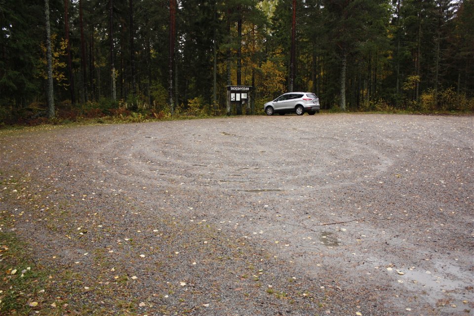 En bil står parkerad på en grusad parkeringsplats vid en skog. Framför bilen finns en informationstavla.