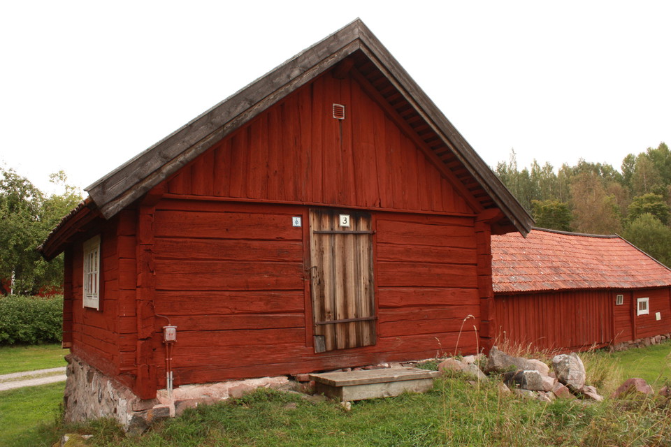 Närbild på en äldre stuga av trä. Ett trappsteg leder upp till dörren in till stugan.