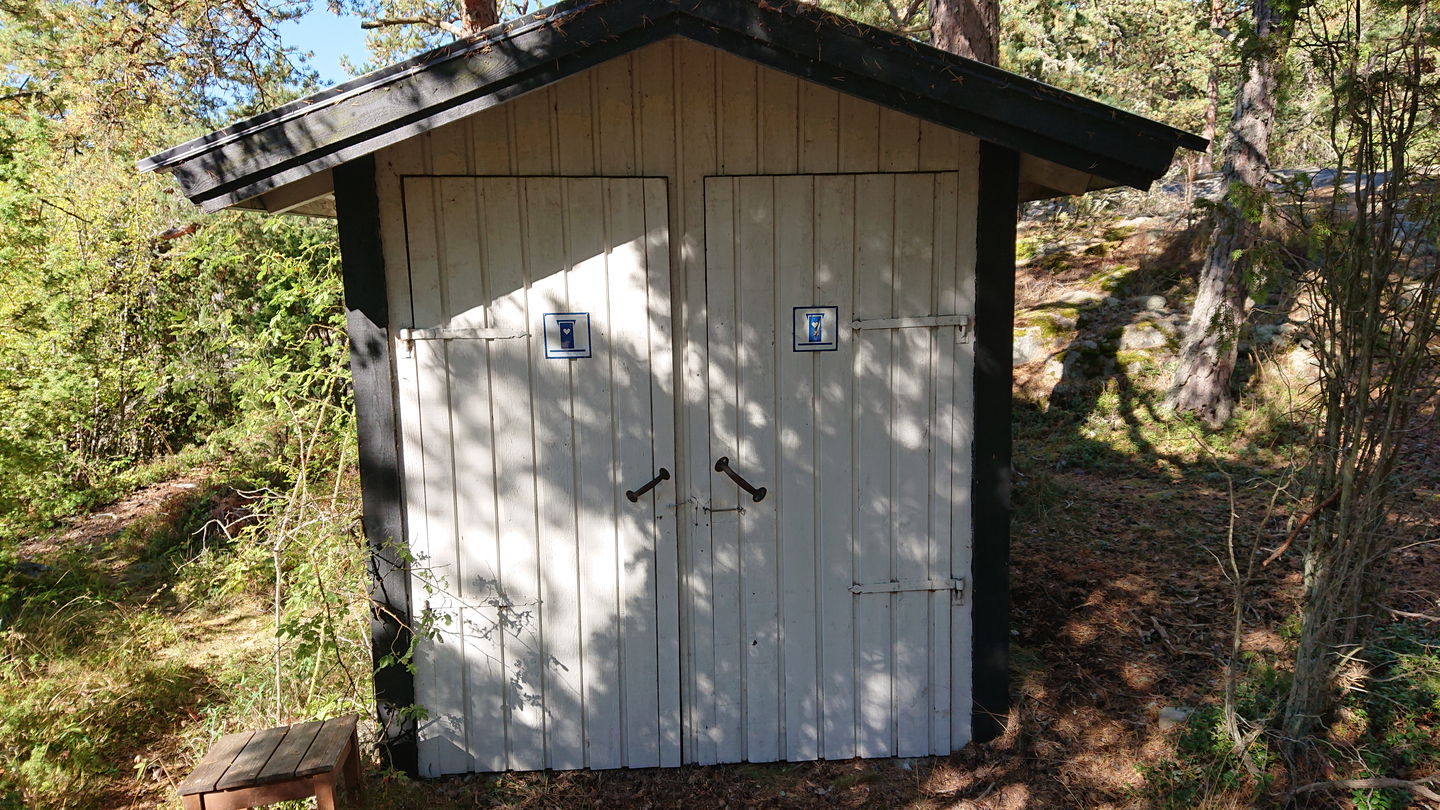 En liten toalettbyggnad med två dörrar står i skogen.