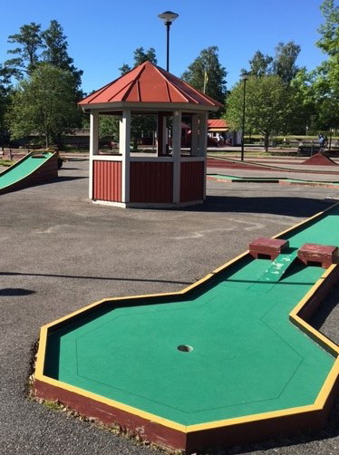Minigolfbana med röda sarger och grön mattbeklädnad, mitt i bilden finns en liten röd byggnad