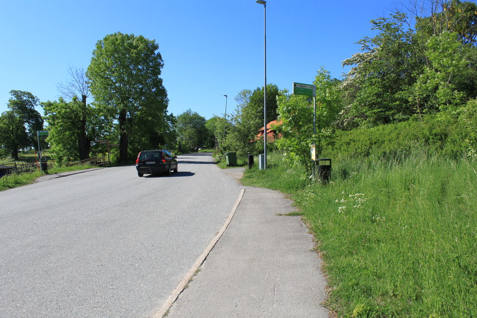 Vid en asfalterad bilväg finns en busshållplats. En bil åker på vägen och det är mycket grönska runtom.