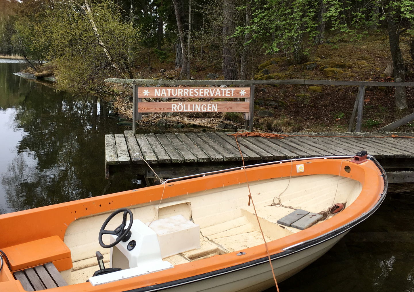 En liten båt står förtöjd vid en träbrygga. På träbryggan finns en skylt med texten "Naturreservatet Röllingen". 