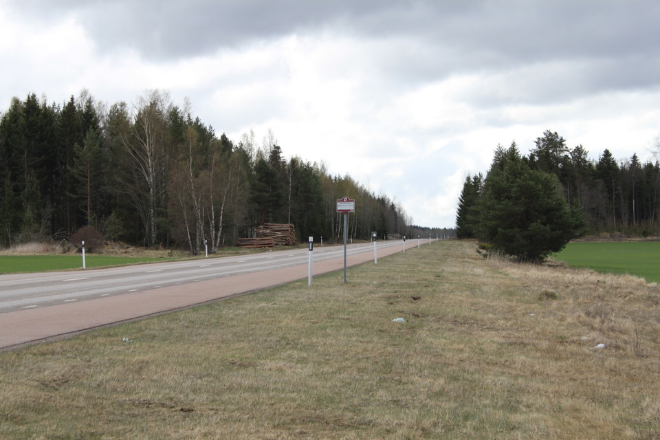 Vid en bilväg står en skylt som markerar busshållsplatsen. I bakgrunden finns skog på båda sidorna av bilvägen.
