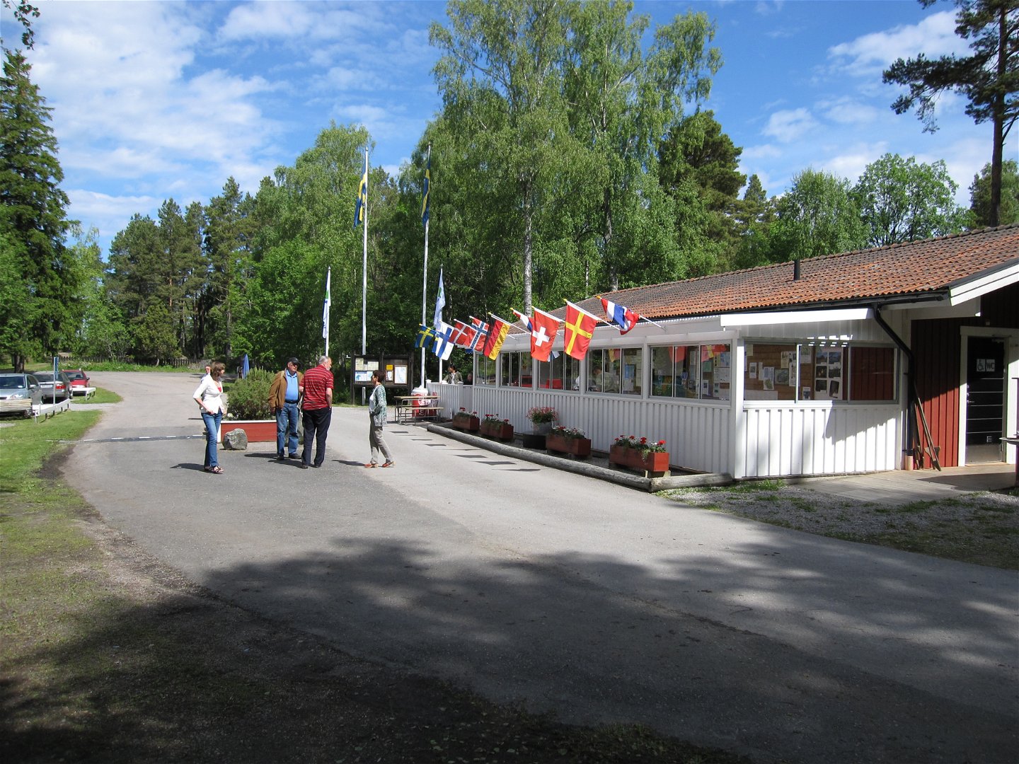 Utanför receptionsbyggnaden står fyra vuxna på en asfalterad väg. Vid byggnaden finns en informationstavla och bänkbord och många olika flaggor är uppsatta på fasaden.