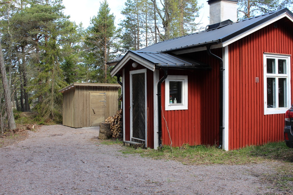 En liten stuga med grusad yta framför. Vid ingången till stugan ligger en hög med ved och i närheten står ett utedass.