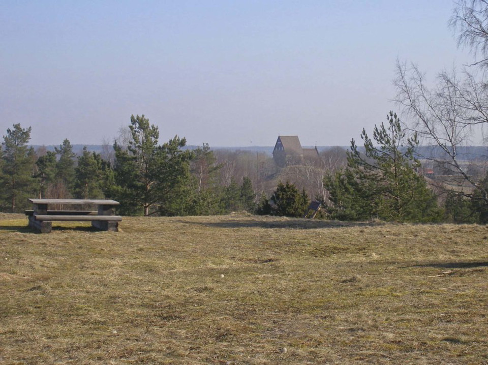 Ett bänkbord står på öppen gräsyta på en höjd. Härifrån har man vy över en skog och i bakgrunden skymtar en hög byggnad.