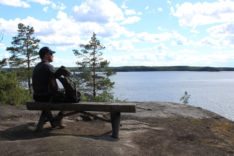 En mat sitter på en sittbänk på ett berg. En ryggsäck lutar mot mannen. Från platsen har man utsikt över vattnet.
