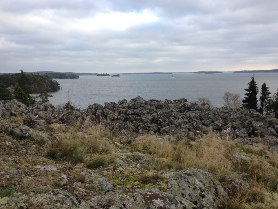 På en hög klippa vid en sjö ligger en hög med större stenar. Från platsen har man utsikt över skärgård och skog.