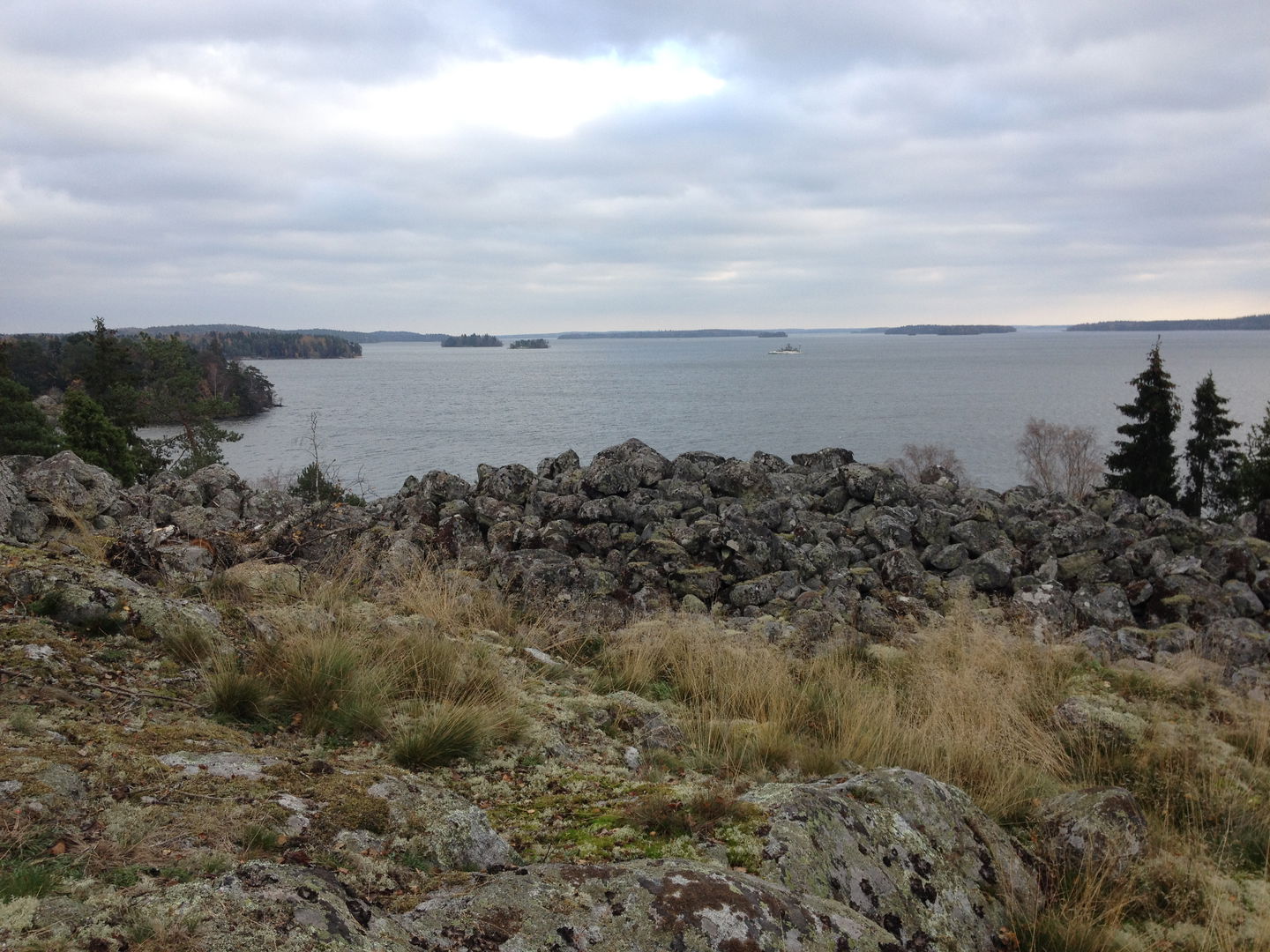 På en hög klippa vid en sjö ligger en hög med större stenar. Från platsen har man utsikt över skärgård och skog.