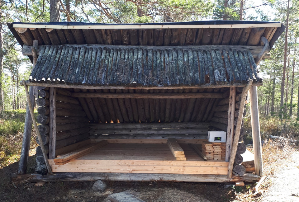 Foto på ett vindskydd rakt framifrån. Virket är gammalt, förutom ett nylagt brädgolv.