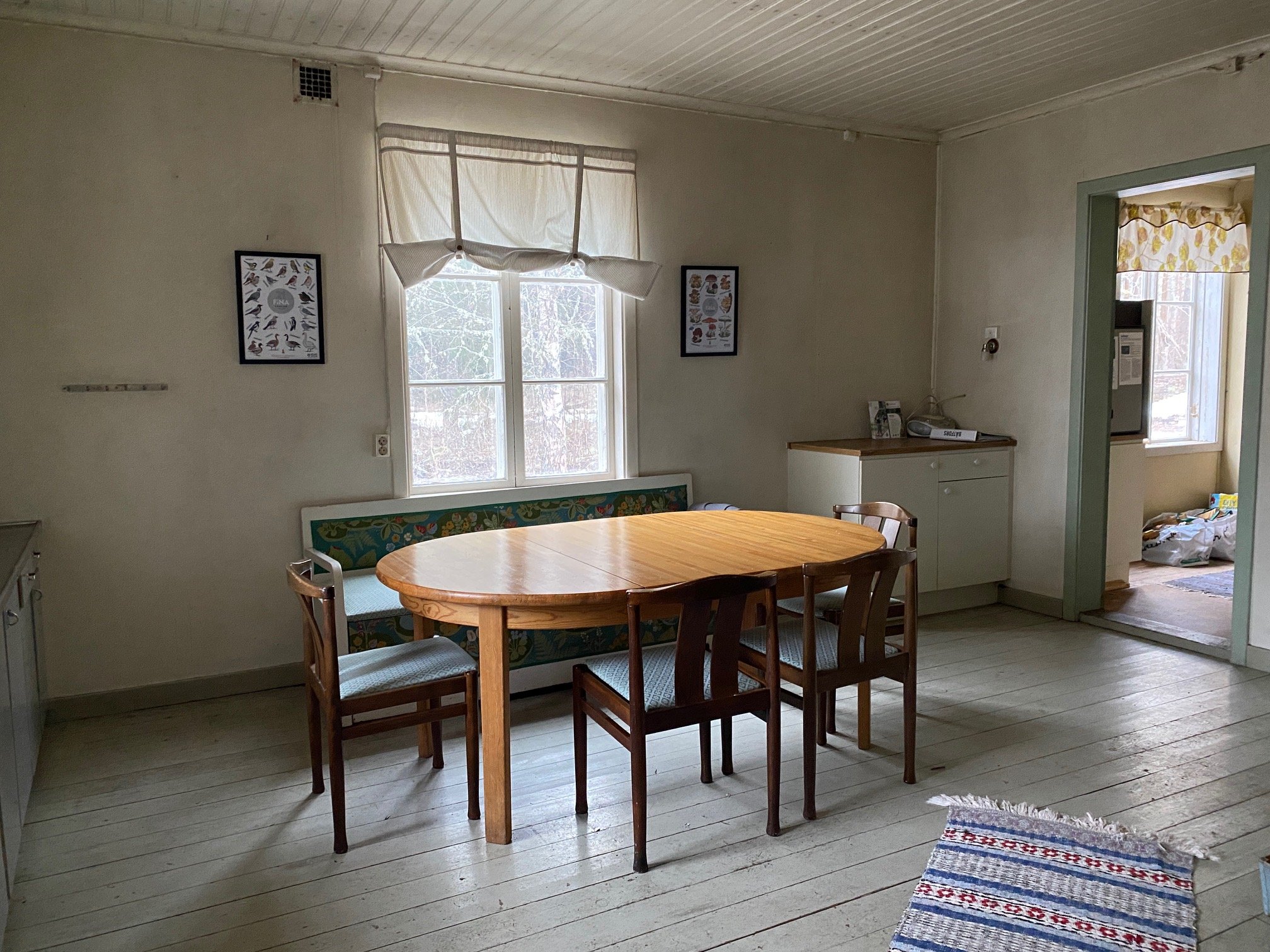 Ett kök inne i ett äldre torp. Ett bord med stolar står mitt i rummet, på golvet ligger en trasmatta och det finns ett par köksbänkar i rummet. På ena väggen hänger två tavlor.