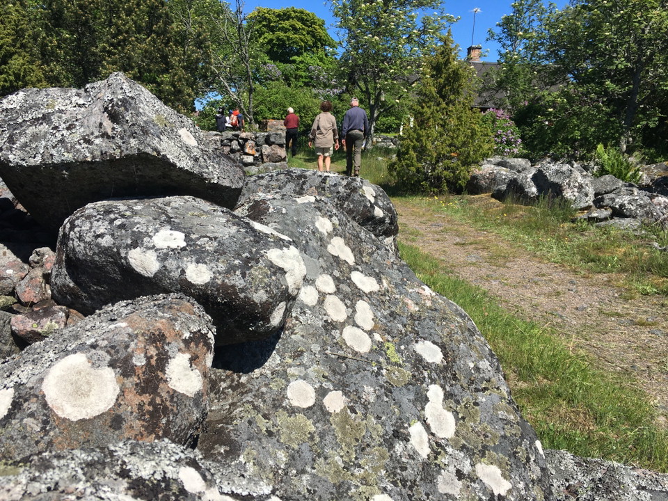 Några personer promenerar längs en stig mellan gärdsgårdar av sten. I bakgrunden finns många träd och mellan dem syns ett hustak med en skorsten. 