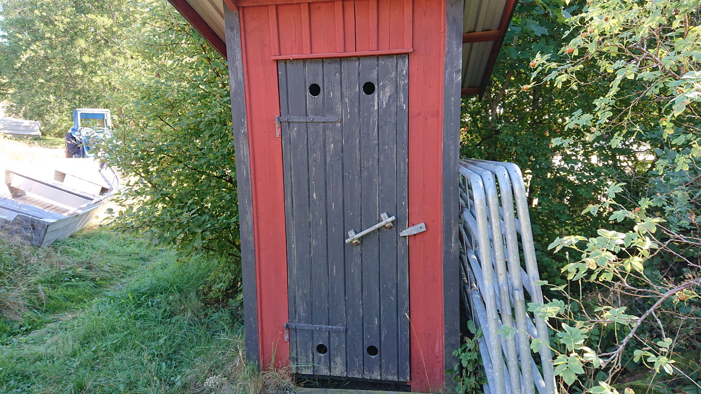 En liten torrtoalett står framför buskage. Några metallstängsel lutar mot ena sidan av toaletten.