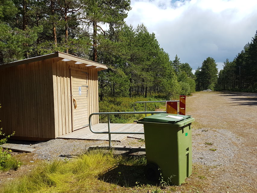 På ena sidan av en stor grusad parkeringsplats står en tillgänglighetsanpassad torrtoalett. En ramp leder upp till toaletten och på ena sidan står en soptunna.