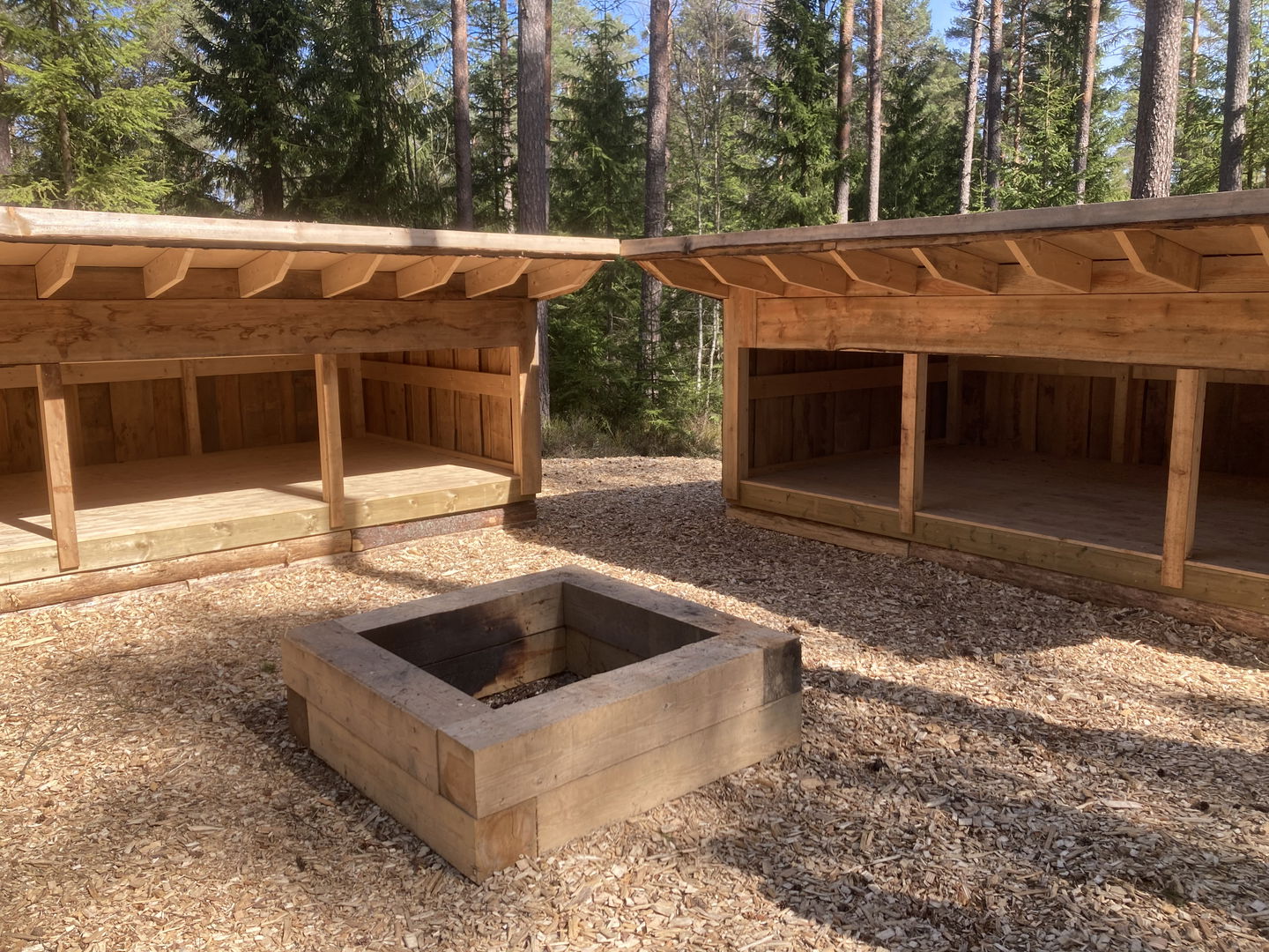 Två vindskydd och en grillplats med skog i bakgrunden