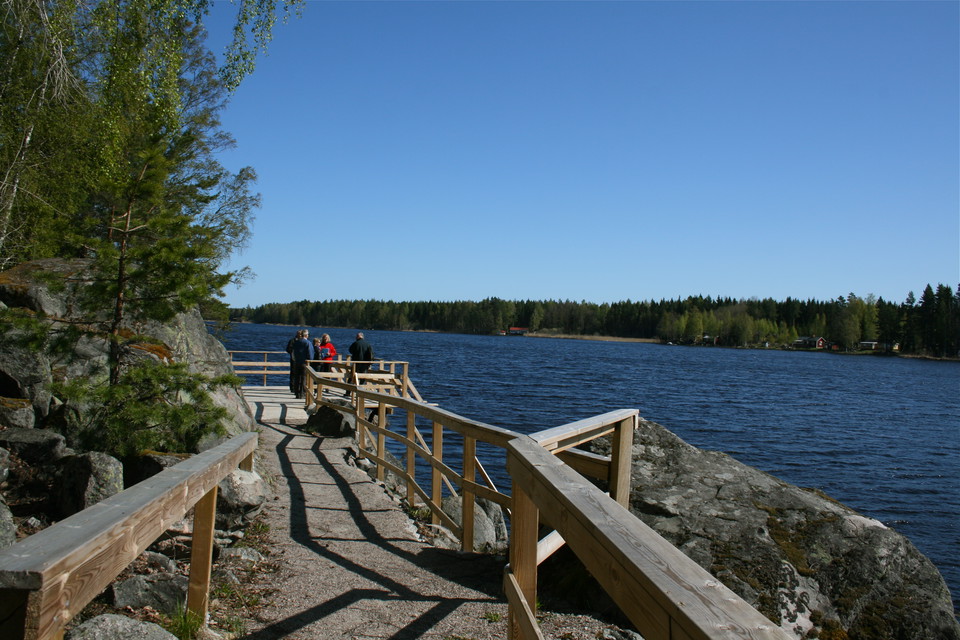 En grusad stig med handräcken på båda sidorna leder fram till en brygga vid vattnet. På bryggan står flera personer och tittar ut över vattnet. 