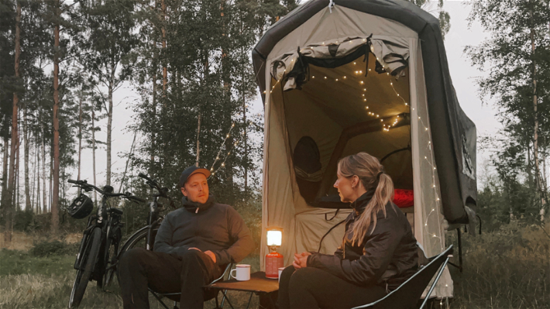 Två personer sitter vid campingbord bredvid tältvagn.