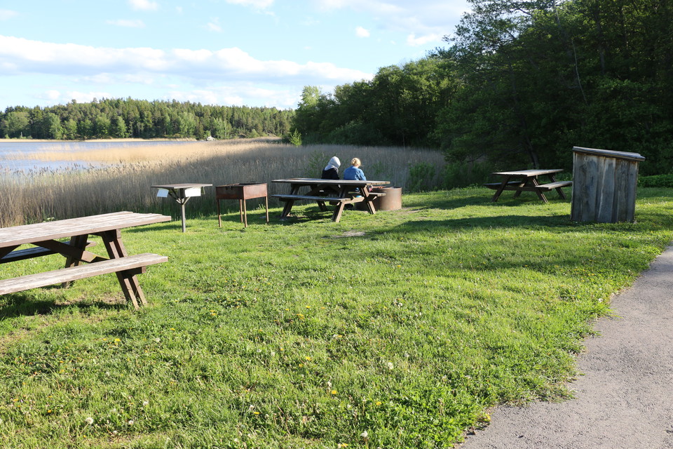 Rastplats intill vattnet med bord och bänkar, grillplats och vedförråd.