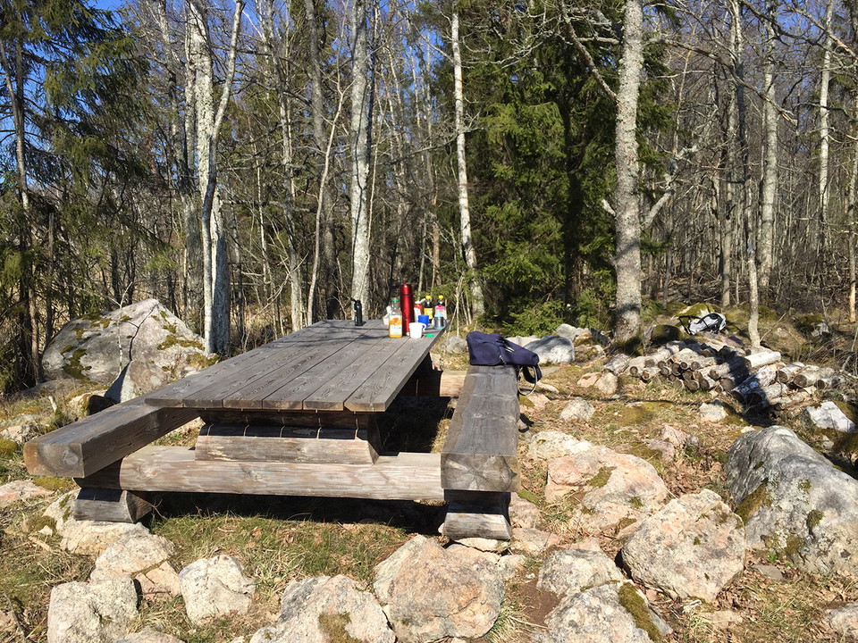 Träbord och träbänkar. En ryggsäck ligger på en av bänkarna. På bordet en termos och läskflaskor.