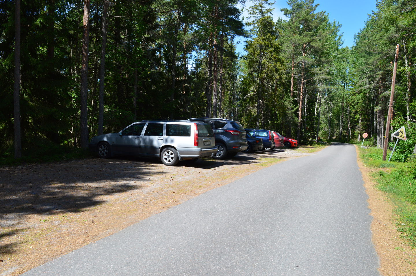 Längs sidan av en asfalterad väg står flera bilar parkerade på en avlång parkeringsplats. Underlaget är jämnt hårdpackat grus.