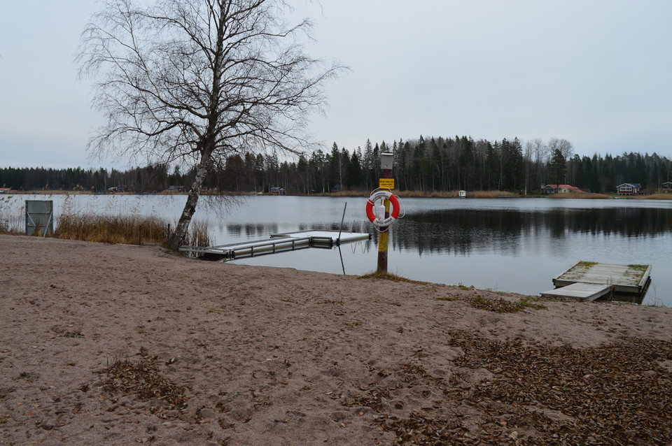 Två flytbryggor ligger i vattnet vid en sandstrand. Vid strandkanten står en stolpe med en livboj på.