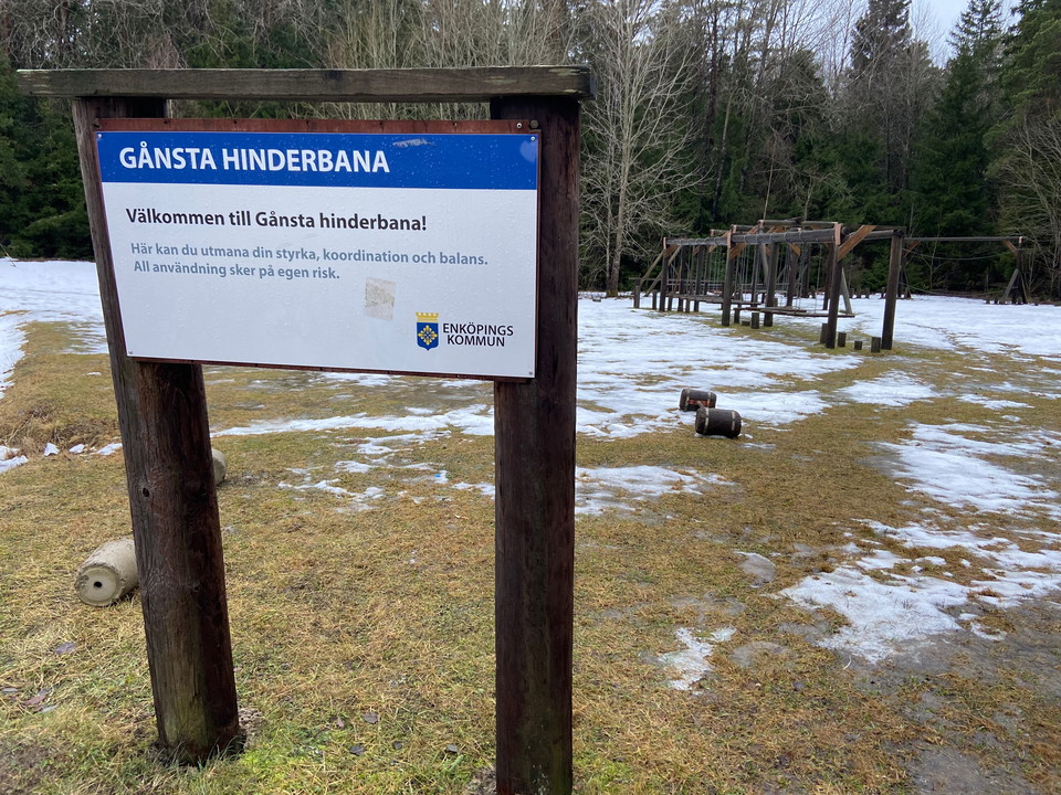 I förgrunden syns en skylt med rubriken Gånsta hinderbana. I bakgrunden syns delar av hinderbanan. 