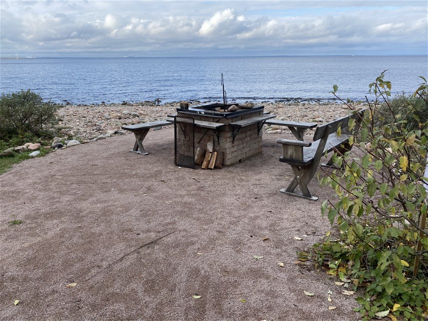 På en jämn, grusad yta vid havet står en grillplats och flera sittbänkar.