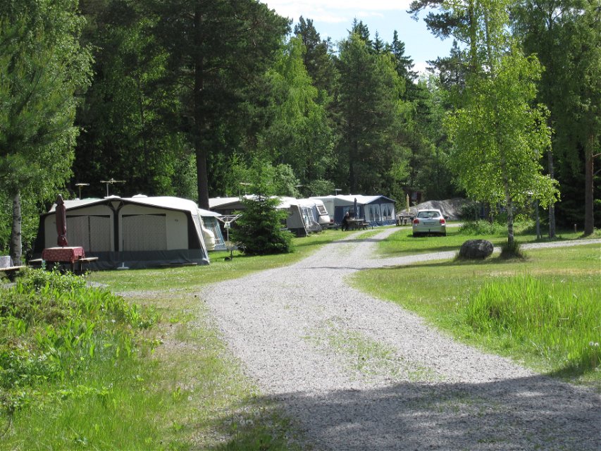 En bred grusväg leder fram till en stor campingplats med flera stora tält och husvagnar parkerade i en rad. Runtomkring är det gott om gröna ytor och träd.