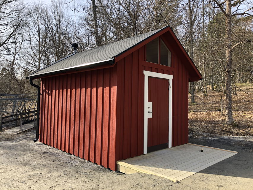 En bred ramp leder upp till en torrtoalett som står på en grusplan. 