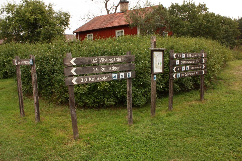 Framför en buske  i närheten av lägergården står en informationsskylt och flera vägvisare som pekar i olika riktningar.