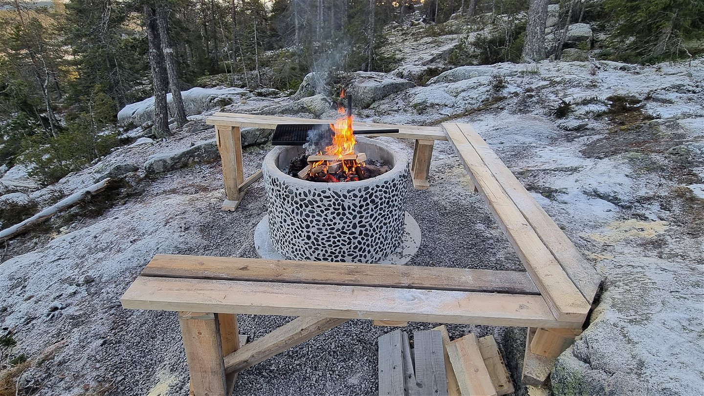 A fireplace with benches on a rocky surface surrounded by coniferous forest. 