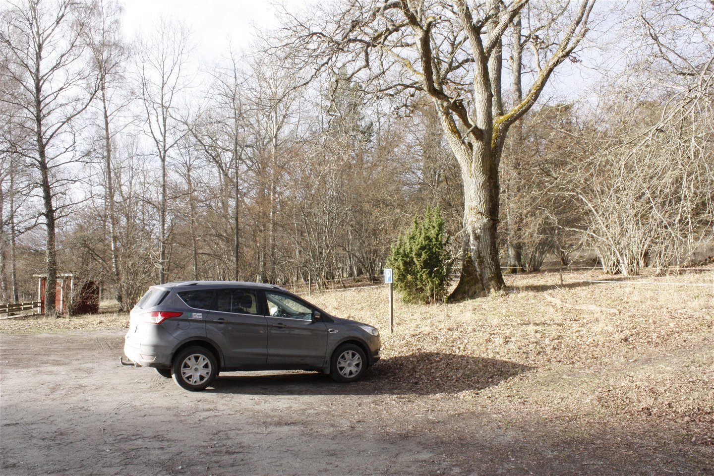 En bil står på en stir grusad parkeringsplats. I bakgrunden syns stora träd och en torrtoalett.