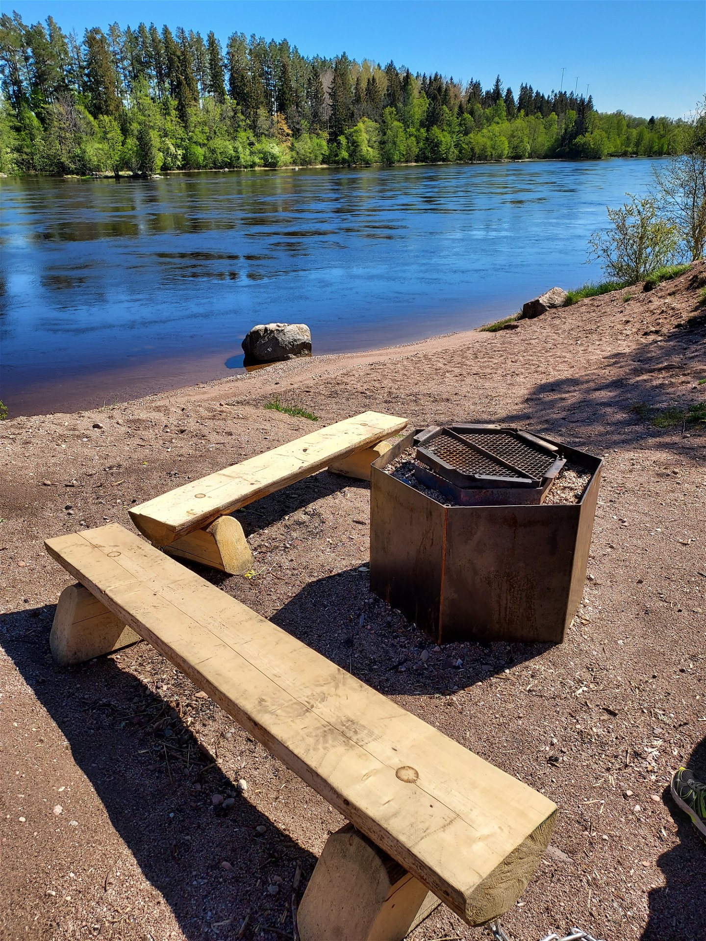 På en sandstrand vid Dalälven står en liten grillplats med två sittbänkar bredvid.