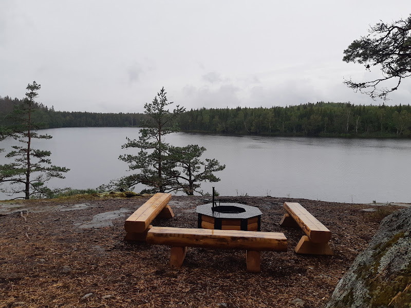Tre träbänkar står runt en eldstad av trä med smideskant. Platsen ligger ganska högt med utsikt över en sjö.