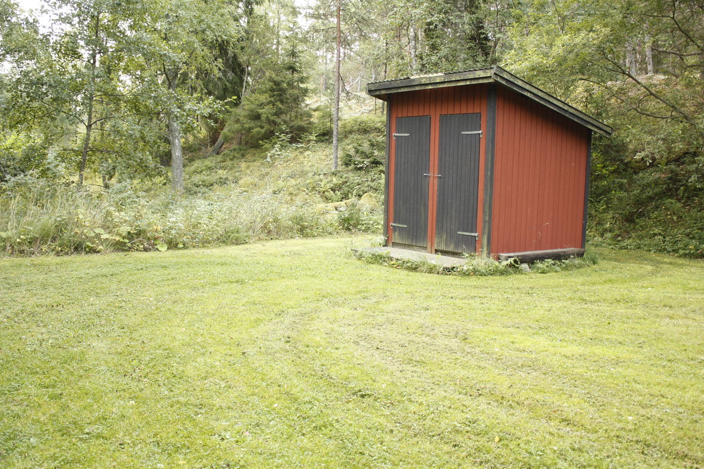 En liten toalettbyggnad med två ingångar står på en gräsplan. I bakgrunden är det skog.