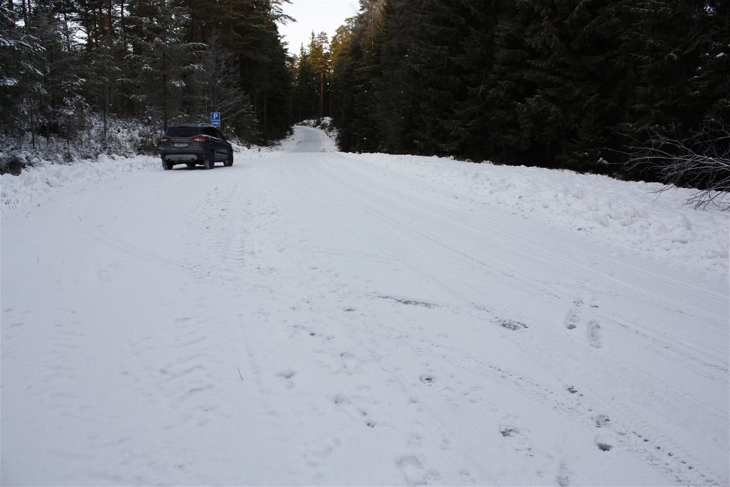 Vid en smal skogsväg finns en avlång parkeringsficka. På parkeringen står en bil och det ligger snö på marken.