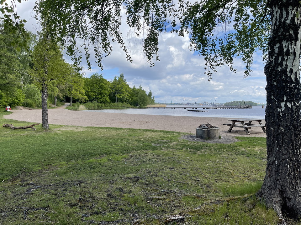 Badplats med sandstrand. En grillstad och ett picknickbord. På avstånd en båtbrygga.