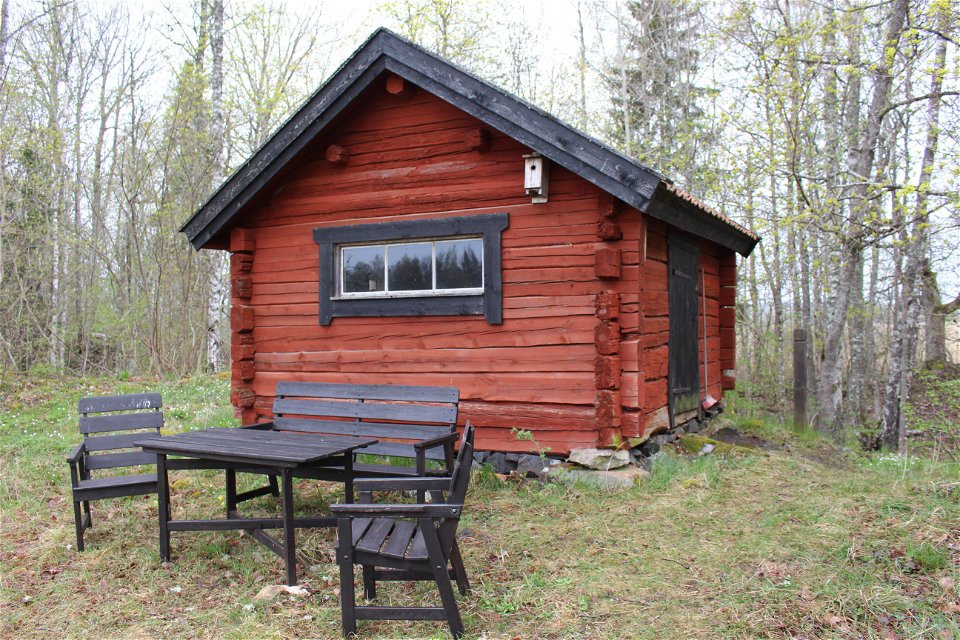 I en skogsglänta står ett litet trähus med ett fönster. Framför huset står ett bord med stolar och en sittbänk runtom sig.