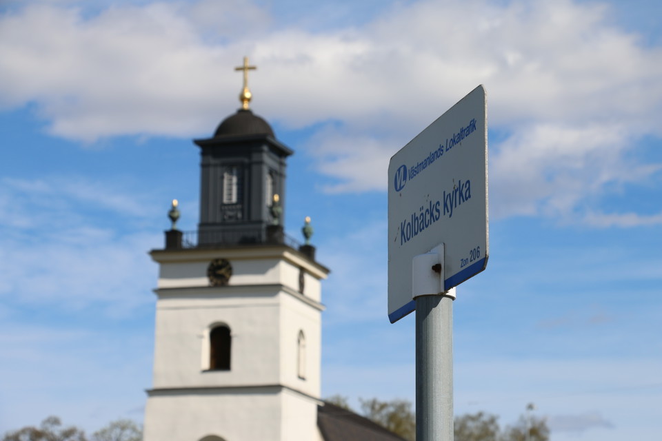 Busshållplatsskylt i närbild med kyrkan i bakgrunden.