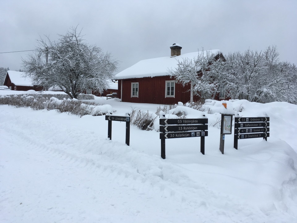 Vid en parkeringsplats står informationsskylt och flera vägvisare. I bakgrunden står flera stugor och det ligger mycket snö på marken.