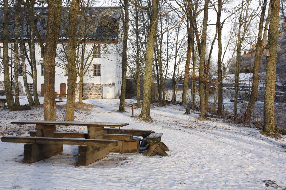 Ett bänkbord och en grillplats står i en dunge med träd runtom. Det ligger snö på marken och i bakgrunden står en byggnad.