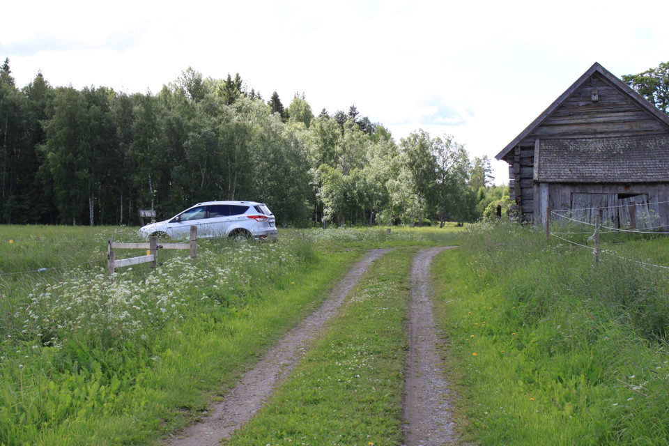 Vid en skogsbilväg finns en liten parkeringsplats med en bil på. Mitt emot bilen står en gammal lada.