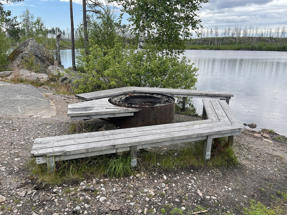 Eldstad med grånade träbänkar vid vatten.