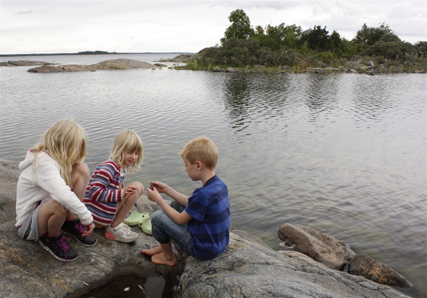Tre barn sitter på en klipphäll och undersöker något som en av barnen håller i handen. I bakgrunden syns havet och en vik med buskar på.